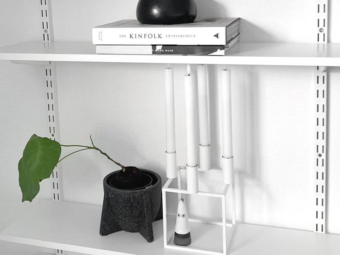 En minimalistisk hvid hylde med bøger, en sort vase, en potteplante med et enkelt blad og en moderne hvid lysestage.