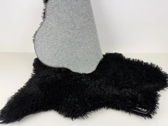 Et sort, loddent tæppe med en grå, mønstret underside, der er delvist løftet, så man kan se dets struktur og et lille sort mærke i hjørnet.