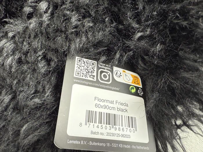 Nærbillede af en sort, struktureret gulvmåtte med en produktetiket med teksten "Floormat Frieda 60x90cm black" og en stregkode, QR-kode og virksomhedsoplysninger.