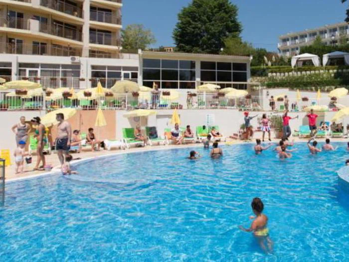 Folk nyder en solrig dag ved et hotels udendørs pool; nogle svømmer, mens andre slapper af på liggestolene med gule parasoller i baggrunden.