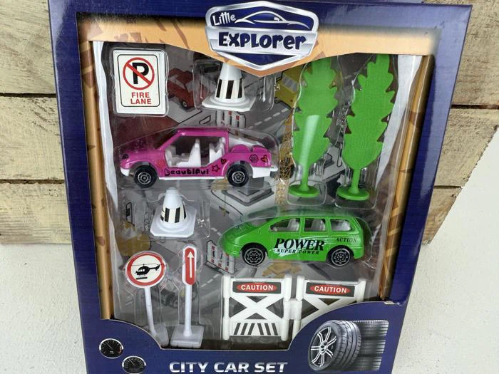 Legetøjsbybilsæt i emballage med en pink og en grøn bil, trafikskilte, træer, kegler og vejafspærringer med påskriften "Caution". På emballagen står der "Little Explorer" og "City Car Set".