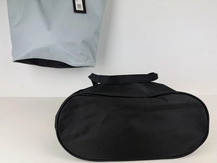 En sort oval taske med hank står på en hvid flade, og bag den hænger en grå taske på væggen.