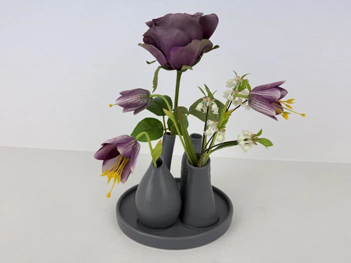Et gråt vasesæt med kunstige lilla og hvide blomster står på en rund bakke mod en almindelig hvid baggrund.
