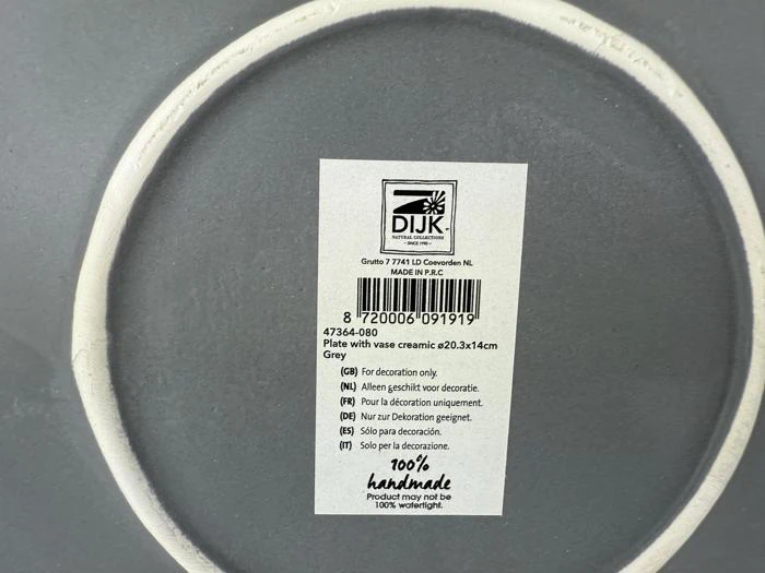 Nærbillede af en produktetiket i bunden af en grå tallerken med mærket DIJK, stregkode, plejeanvisninger og en bemærkning om, at den kun er til pynt og håndlavet i Kina.