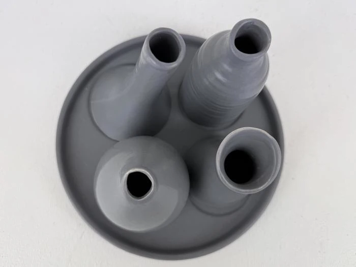 Fra oven ses fire grå keramikvaser i forskellige former og størrelser arrangeret på en rund grå bakke mod en hvid baggrund.