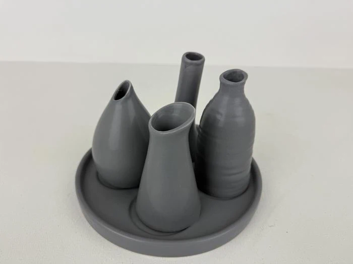 Fire små, grå, forskelligt formede keramikvaser er anbragt lodret på en rund, matchende bakke mod en almindelig hvid baggrund.