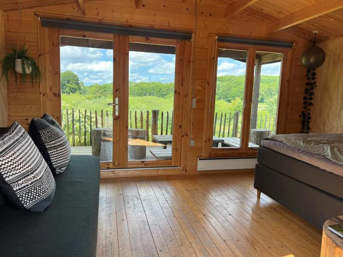 En hyggelig træhytte indvendigt med store vinduer og glasdøre med udsigt over et frodigt grønt landskab; en sort sofa og seng er synlig indeni.