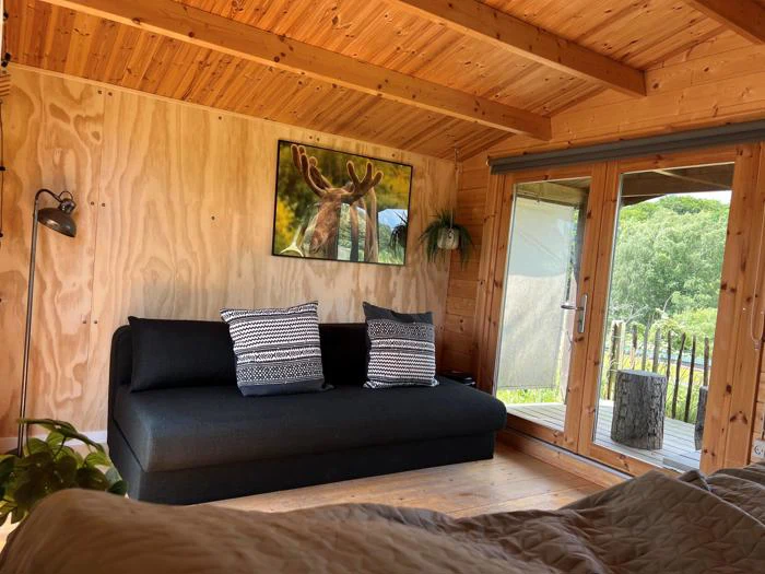 Et hyggeligt træhytteinteriør med en sort sofa, to mønstrede puder, et elgmaleri, store vinduer og udsigt til grønne områder udenfor.