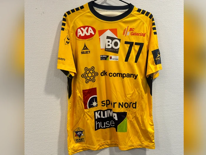 En gul sportstrøje med sorte detaljer, nummer 77 og forskellige sponsorlogoer, der hænger på en sort bøjle mod en hvid struktureret væg.
