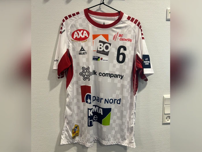 En hvid og rød sportstrøje med forskellige sponsorlogoer, tallet 6 og navnet BO hænger på en sort bøjle mod en lys væg.