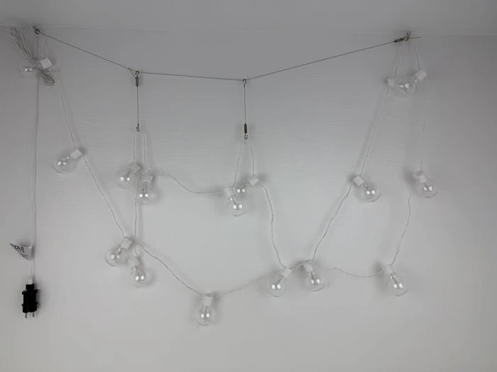 En hvid lyskæde med blottede pærer er draperet i et løst zigzagmønster og fastgjort til en hvid væg. Et sort stik er synligt til venstre.