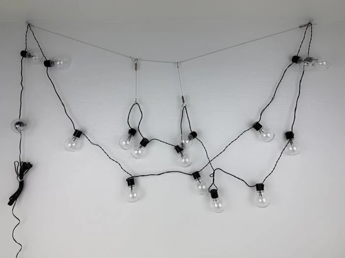 En række klare pærer hænger på en hvid væg, løst draperet i et zigzagmønster med sorte snore.