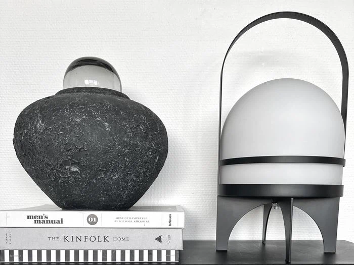 En struktureret sort vase med en glaskugle på toppen står på stablede bøger ved siden af en moderne, æggeformet lampe med en sort metalramme.