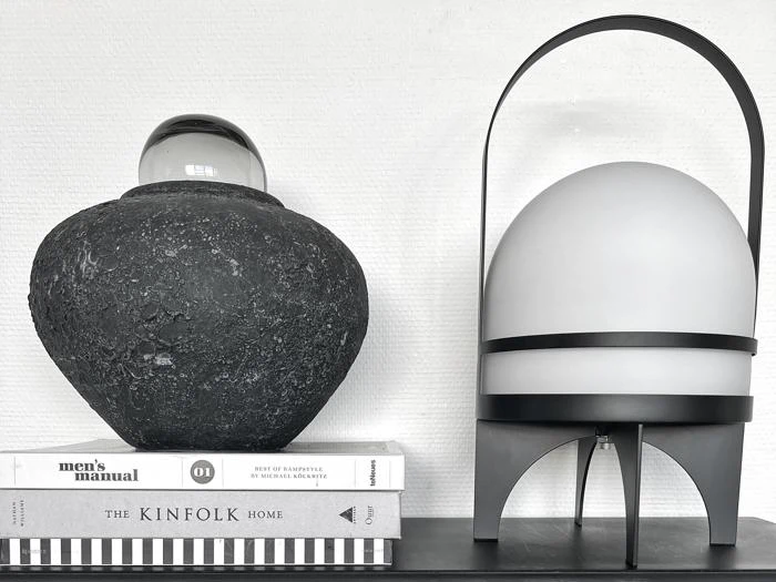En sort tekstureret vase med en glaskugle på toppen står på stablede bøger ved siden af en moderne hvid lampe med en sort ramme, alt sammen mod en hvid væg.