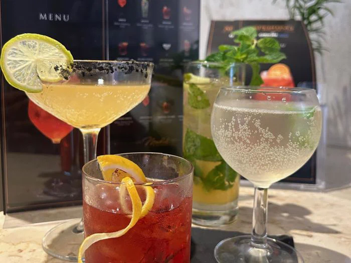 Fire forskellige cocktails garneret med lime, mynte og citronskal står på et bord, og drinkskortet er synligt i den slørede baggrund.