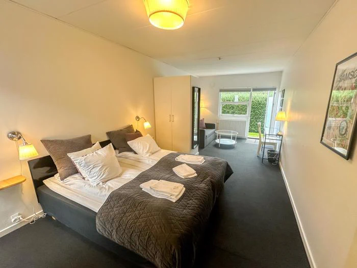 Et lyst hotelværelse med to enkeltsenge skubbet sammen, et klædeskab, en sofa, et skrivebord og et vindue med udsigt til grønne områder. Håndklæderne ligger pænt på sengene.