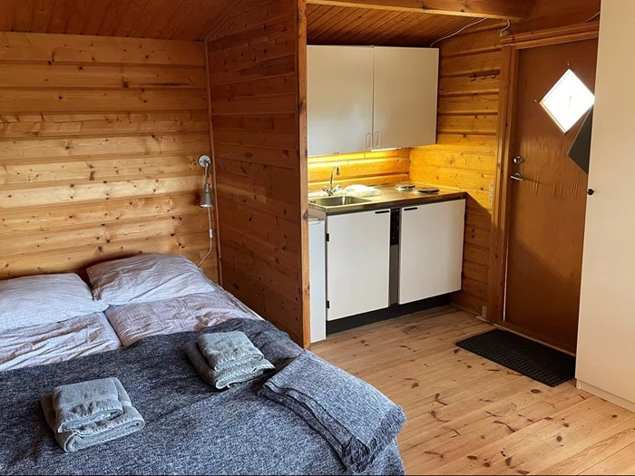 En hyggelig træhytte indrettet med en seng, foldede håndklæder, et lille tekøkken med vask og komfur og en trædør med et diamantformet vindue.