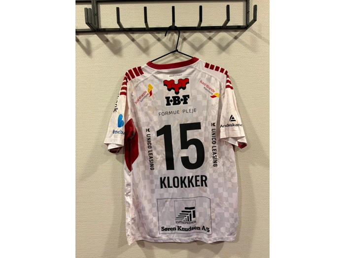 En hvid og rød sportstrøje med nummer 15 og navnet KLOKKER hænger på en vægkrog. På ryggen af trøjen ses forskellige sponsorlogoer.