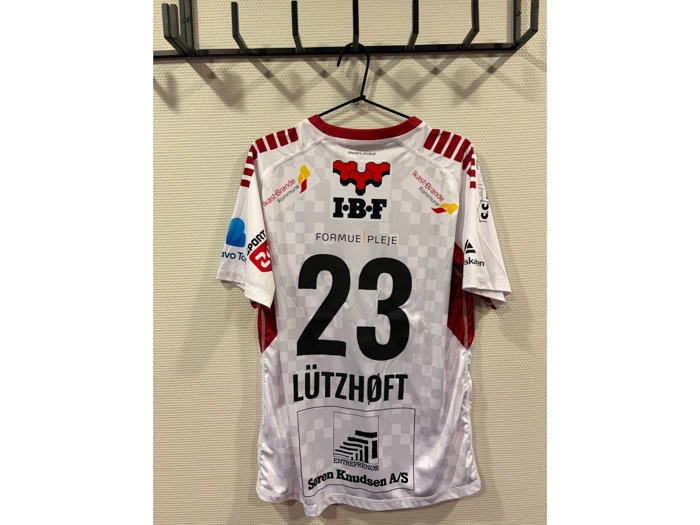 En hvid og rød sportstrøje med nummer 23 og navnet Lützhøft hænger på en vægkrog og er prydet med forskellige sponsorlogoer.