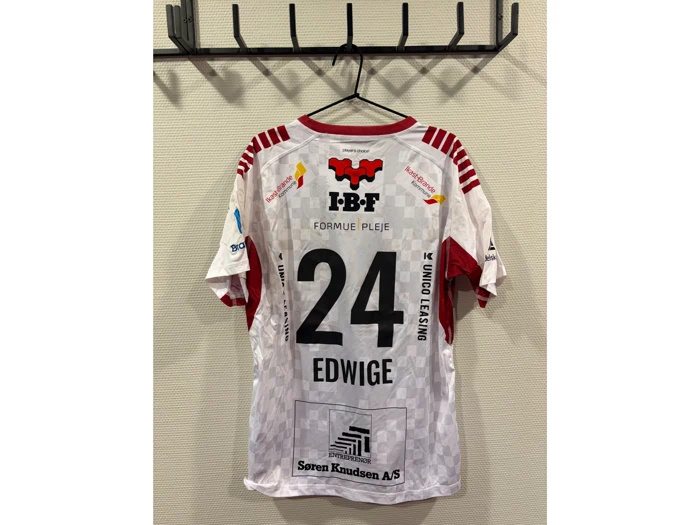 En hvid og rød sportstrøje med nummer 24 og navnet Edwige hænger på en sort vægkrog. På ryggen ses forskellige sponsorlogoer.