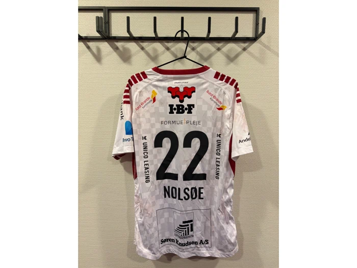 En hvid sportstrøje med røde detaljer hænger på en vægkrog. På ryggen ses nummeret 22 og navnet NOLSØE samt forskellige sponsorlogoer.