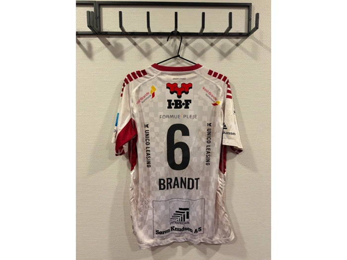 En hvid og rød sportstrøje med nummer 6 og »BRANDT« på ryggen hænger på en vægmonteret frakkestang. Der ses flere sponsorlogoer på trøjen.