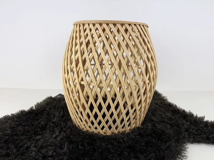 En vævet, lys rattan-kurv med et åbent mønster på kryds og tværs sidder på en struktureret sort overflade mod en almindelig hvid baggrund.