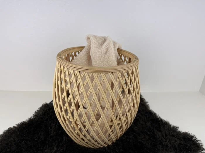 Et beige håndklæde, der er draperet i en rund, åben flettet kurv, placeret på et sort, loddent tæppe mod en hvid baggrund.