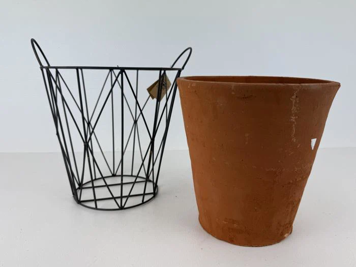 En sort trådkurv og en terracotta-blomsterpotte er placeret side om side på en hvid flade mod en almindelig hvid baggrund.