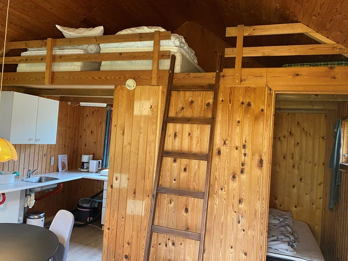 En hyggelig træhytte med en hems, som man får adgang til via en stige, et lille køkkenområde og en anden seng, som er delvis synlig i en lavere krog.