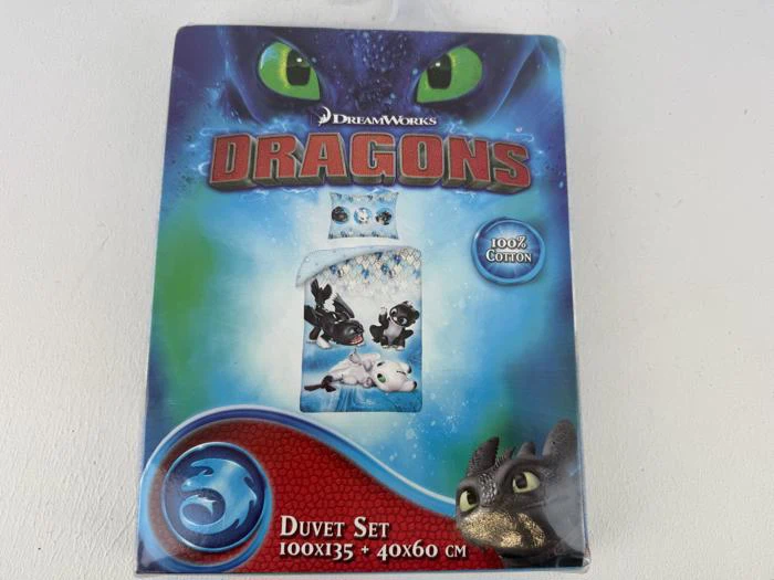 Æske med DreamWorks Dragons dynesæt med Tandløs og andre dragefigurer, mærket 100 % bomuld, størrelse 100x135 + 40x60 cm, med blåt og rødt design.