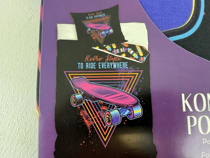 Sengesæt med et neonfarvet skateboard og retrotrekanter, teksten Retro Style TO RIDE EVERYWHERE på dynen og matchende pudebetræk, alt sammen på en lilla baggrund.