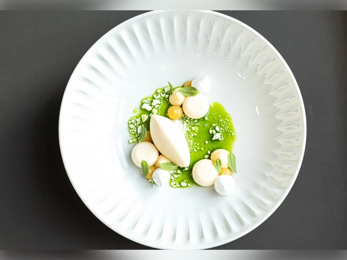 En hvid tallerken med en moderne dessert: grøn sauce, klatter af fløde, små marengs, mynteblade og en oval kugle is, kunstnerisk arrangeret.