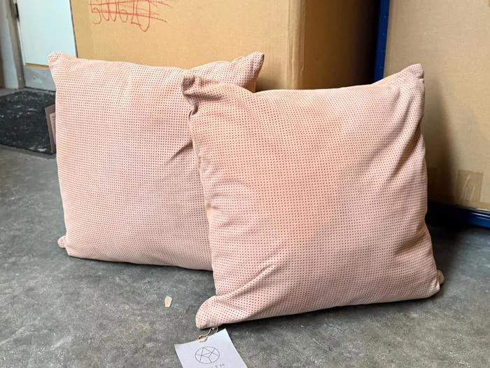 To lyserøde, firkantede puder med diskrete prikmønstre ligger på et gråt betongulv foran store brune papkasser.