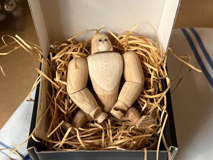 En lille gorillafigur af træ ligger i halm som fyldmateriale inde i en åben kasse på et bord med et stribet klæde.