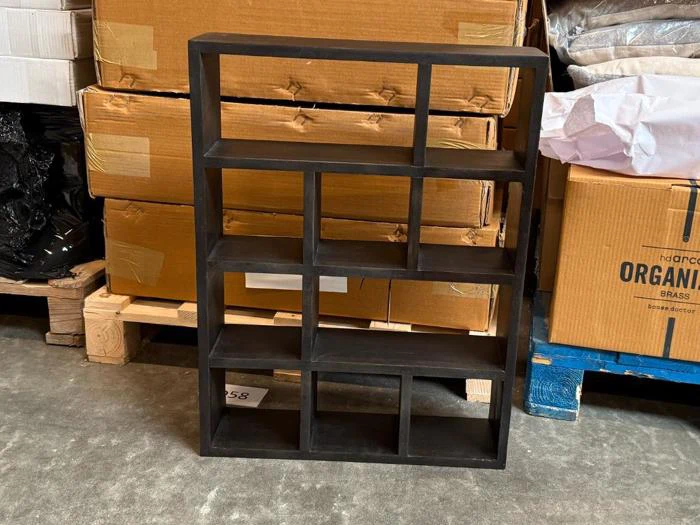 En sort hylde med flere rum står på et betongulv, omgivet af stablede papkasser og palleter indpakket i plast i et lagerlokale.