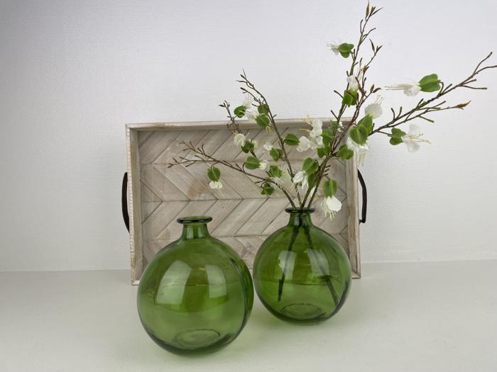 To runde grønne glasvaser, den ene med hvide og grønne kunstige blomster, står foran en træbakke med mørke håndtag mod en almindelig hvid baggrund.