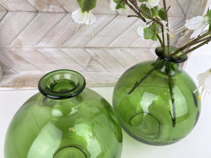 To runde, grønne glasvaser, den ene med hvide blomster og grene, er placeret på en hvid flade foran en træmønstret baggrund.