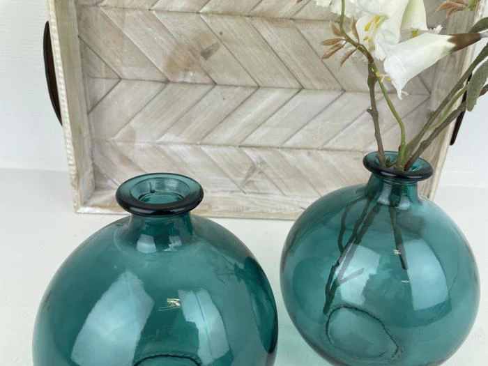 To runde blågrønne glasvaser er placeret foran en rustik træbakke; den ene vase indeholder hvide kunstige blomster.
