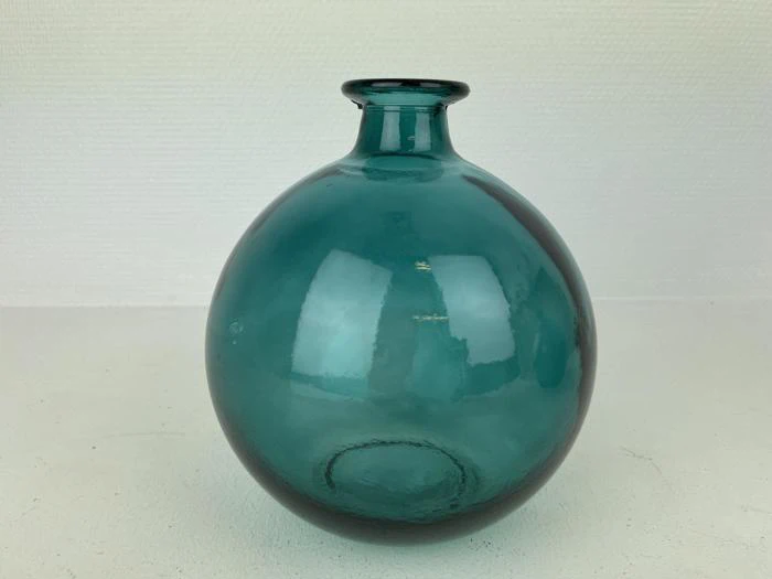 En rund, blågrøn glasvase med en lille, smal hals står på en hvid overflade mod en almindelig, struktureret hvid baggrund.
