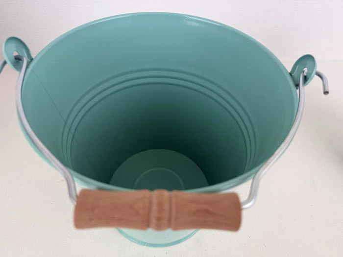 En blågrøn metalspand med træhåndtag, set ovenfra, på en hvid overflade.