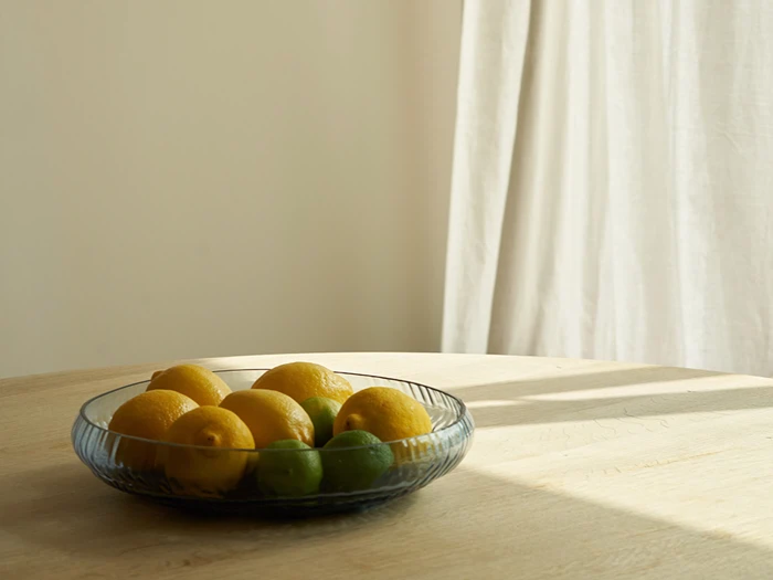 En glasskål fyldt med citroner og limefrugter står på et lyst træbord, hvor sollyset strømmer ind gennem et vindue med hvide gardiner.