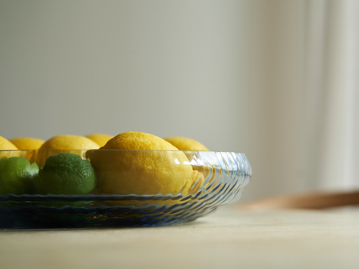 En glasskål med gule citroner og grønne limefrugter står på en lys flade med en blød, neutral baggrund.