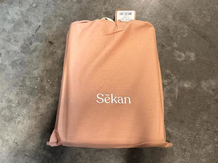 En ferskenfarvet stofpose af mærket Sekan med en mærkeseddel, der ligger på en grå, let plettet betonoverflade.