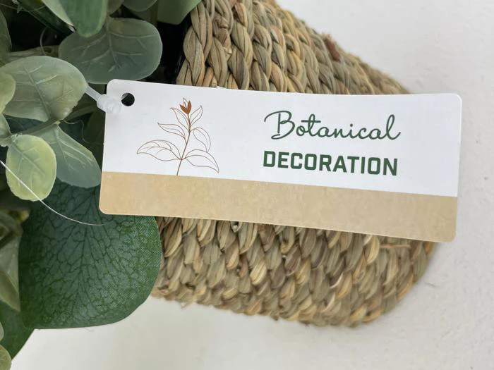 Et mærke med teksten Botanical Decoration sidder på en flettet kurv med grønne kunstige blade, der er delvist synlige omkring den.