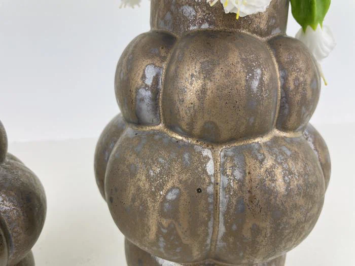 Nærbillede af en struktureret, metallisk bronzevase med et løgformet, segmenteret design med hvide blomster på en ensfarvet baggrund.