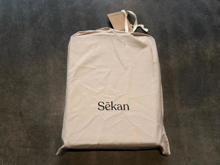 En beige stofpose med ordet »Sēkan« trykt på ligger på en mørk overflade, og der sidder en lille brun etiket fast øverst på den.