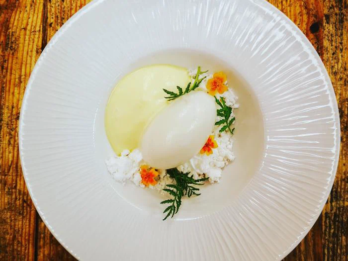 En hvid dessert, muligvis sorbet, serveret med cremet sauce, spiselige blomster og grønne krydderurter på en struktureret hvid tallerken på et rustikt træbord.