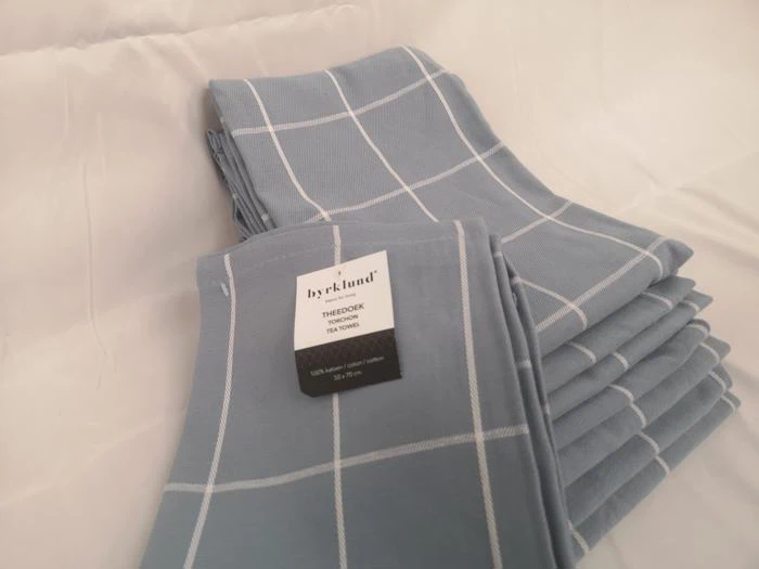 En stak blå viskestykker med et hvidt gittermønster og en synlig etiket med teksten bycklund THEODOR TEA TOWEL.