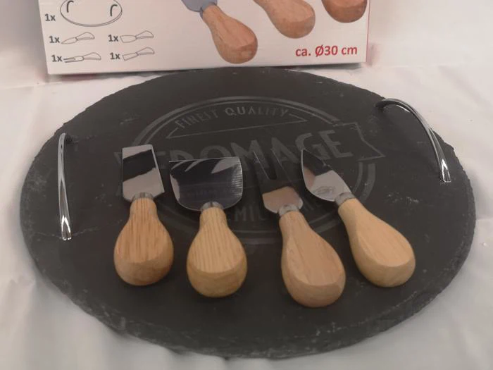 Et rundt ostebræt af sort skifer med metalhåndtag og tre osteknive med træhåndtag arrangeret på det. En produktkasse er delvist synlig i baggrunden.